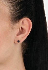 Närbild av ett öra med mörkt hår som är försiktigt bakom, med en liten, rund, fasetterad svart stiftörhänge på örsnibben.