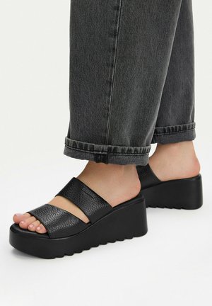 Sandalias negras de plataforma con dos tiras anchas de cuero, usadas con vaqueros gris oscuro remangados y pies descalzos.