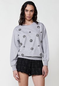Felpa grigia con motivi floreali ricamati, maniche lunghe e colletto rotondo. Abbinata a pantaloni corti neri con una finitura a frange.