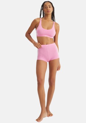 Kobieta stojąca boso, ubrana w różowy sportowy stanik i wysokie szorty z białymi paskami, pozująca z jedną ręką na biodrze.