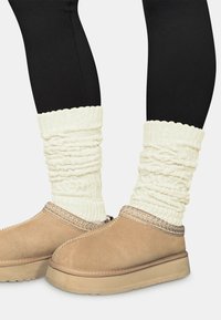 Hellbraune Wildleder-Slipper mit dicker Sohle, ergänzt durch eine strukturierte beige Strick-Stulpe mit geripptem oberen Rand, getragen über schwarzen Leggings.