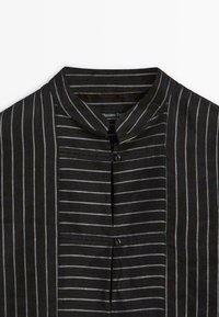 Camicia a righe nere con colletto alla mandarino, chiusura con bottoni e diverse larghezze delle righe in un tessuto setoso. Etichetta visibile all'interno.