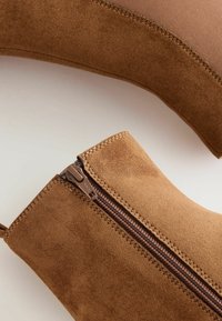 Rudos suede aulinukai iki kulkšnių su šoniniu užtrauktuku, tekstūruota paviršiumi ir minimalistiniu dizainu. Apima detalizuotą siuvinėjimą per siūles.