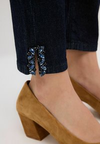 Jeans de mezclilla oscura con una abertura en el dobladillo adornada con embellishments similares a gemas en azul y negro, combinados con zapatos de tacón bloque de gamuza en color marrón.
