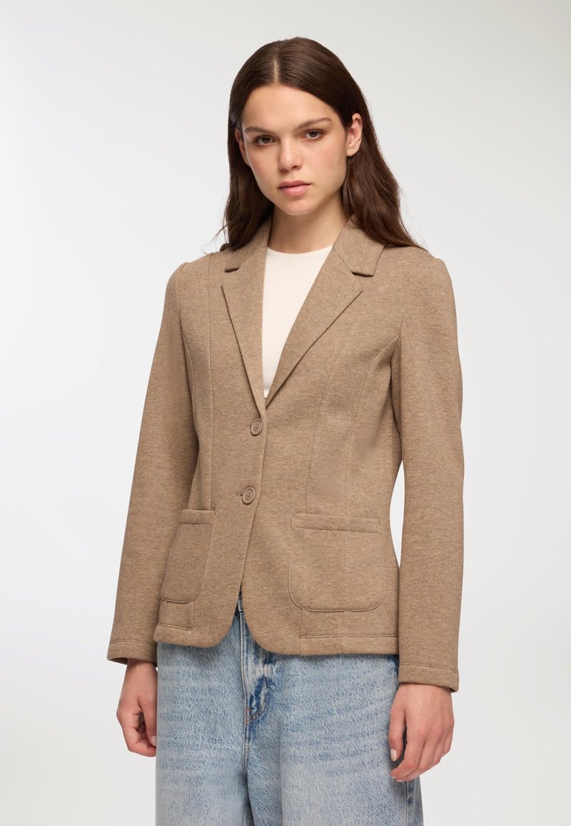 Blazer beige in tessuto testurizzato, vestibilità su misura, chiusura con un bottone, colletto a rever e due tasche frontali. Abbinato a denim chiaro.