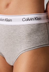 Γκρι εσώρουχο Calvin Klein με λευκή ελαστική μέση, φορεμένο από ένα άτομο που δείχνει μέρος της κάτω κοιλιάς και του άνω μηρού.