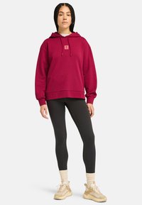 Burgundy huva sweatshirt med en framlogo patch, parat med svarta leggings och beige sneakers. Har en avslappnad passform och ribbade manchetter.