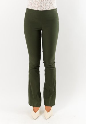 Donna con pantaloni aderenti verde scuro e décolleté bianchi a punta, in piedi davanti a uno sfondo bianco uniforme.