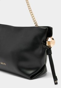 Sort læder skuldertaske med en guld kædestropp og en dekorativ geometrisk charm. Har en glat tekstur og subtilt brand logo.