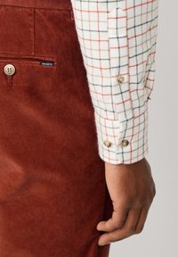 Bruine ribfluwelen broek heeft een zijzak met een knoop en een shirt met een wit-rood ruitpatroon. Details omvatten afgeronde manchetten en twee knopen.