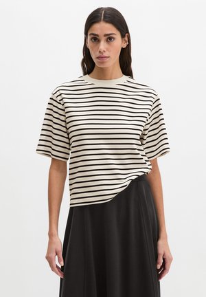 Kobieta ubrana w kremową i czarną bluzkę w poziome paski z krótkim rękawem, włożoną do wysokiej czarnej spódnicy, na jednolitym tle.