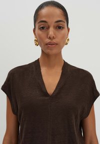 Top marrone con scollo a V realizzato in un tessuto testurizzato, con maniche corte e una vestibilità ampia. Orecchini circolari dorati di grandi dimensioni completano il look.