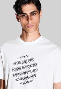 T-shirt bianco a maniche corte con una grafica circolare contenente nomi di città in testo nero. Tessuto di cotone liscio con una vestibilità rilassata.