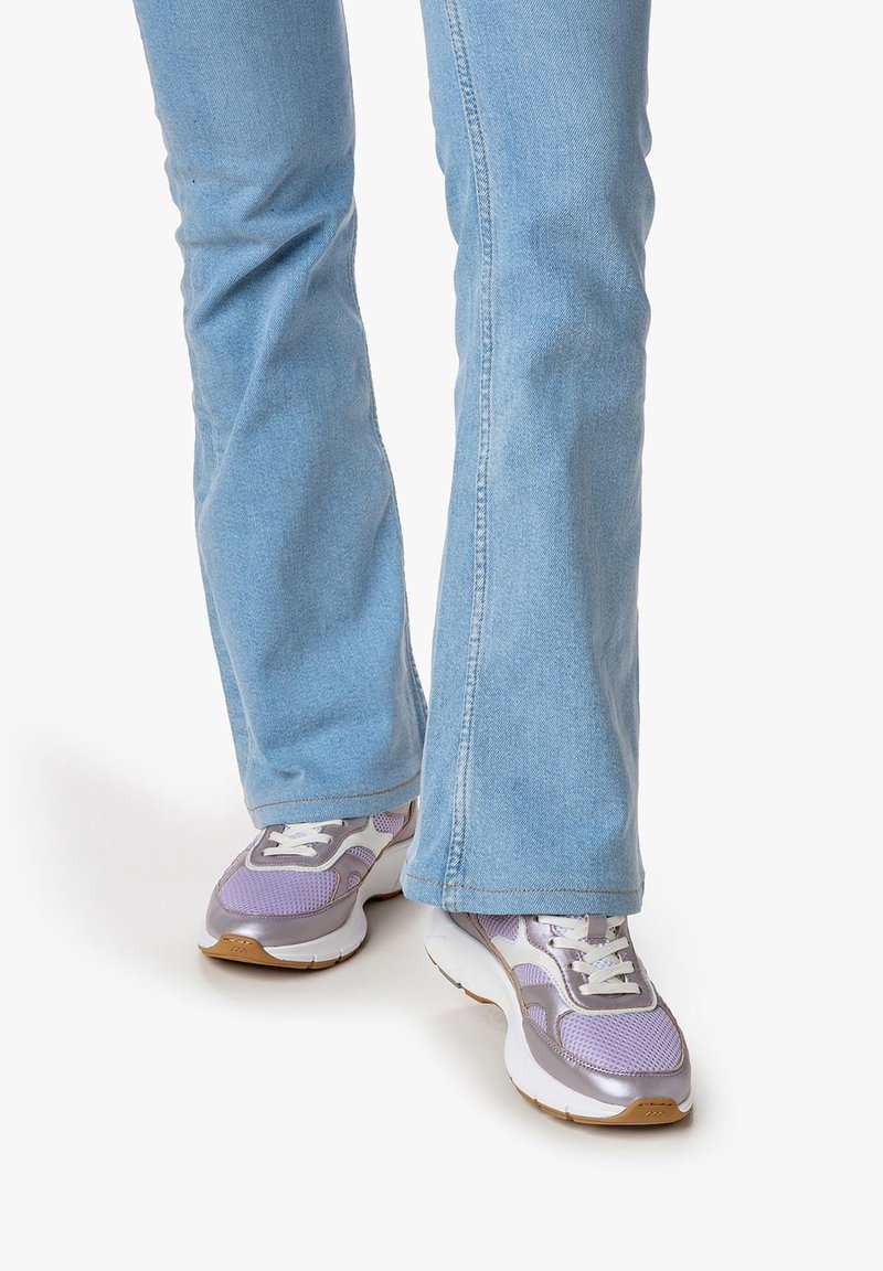 Jasnoniebieskie dżinsy o rozszerzanej nogawce w połączeniu z sportowymi fioletowo-białymi trampkami, które mają siatkowe i gładkie akcenty, na jednolitym tle.