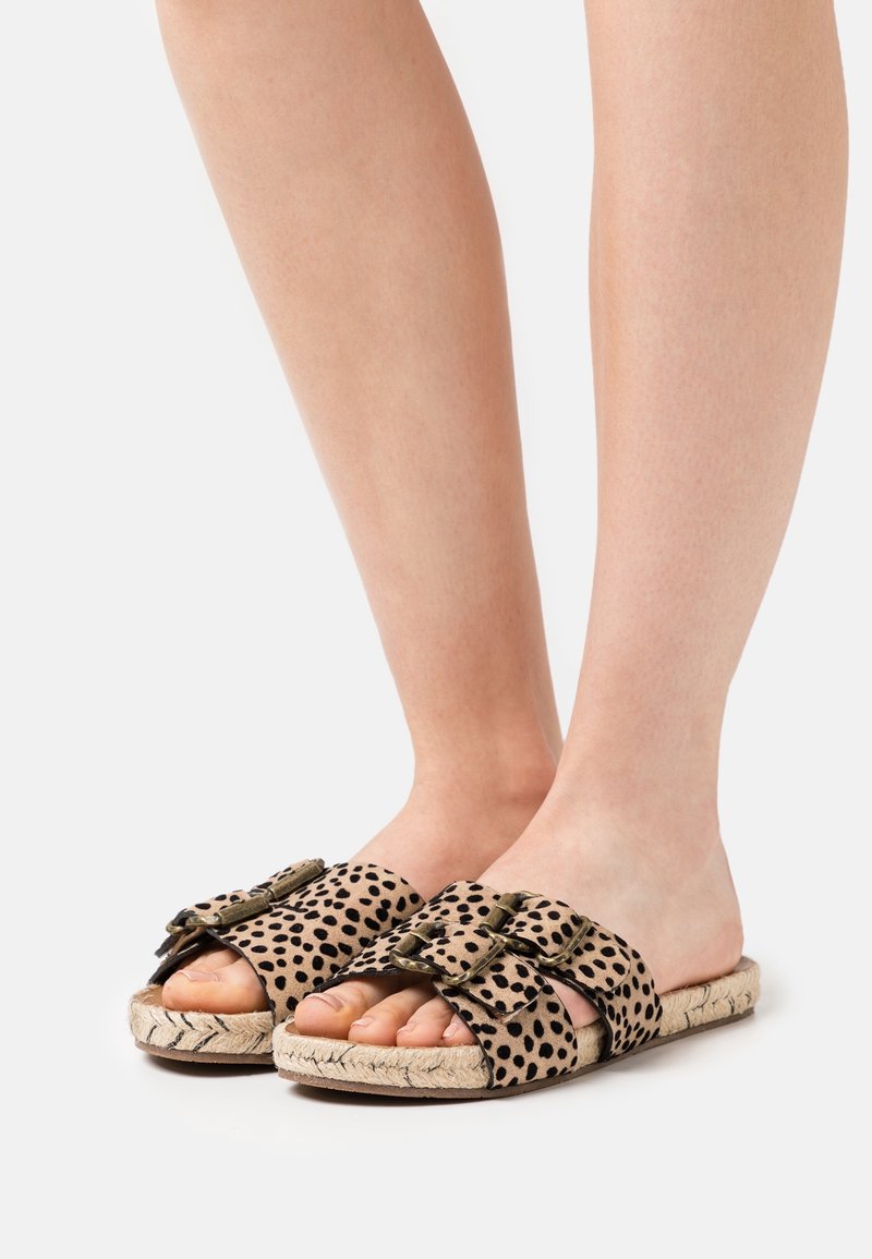 Pieds portant des sandales plates à bout ouvert avec des brides beige et noires à taches et deux boucles métalliques sur des semelles tissées de couleur claire.