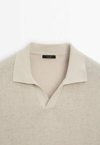 Polo en maille beige avec un col côtelé et une ouverture en V, montrant une étiquette de marque noire à l'intérieur de l'encolure.