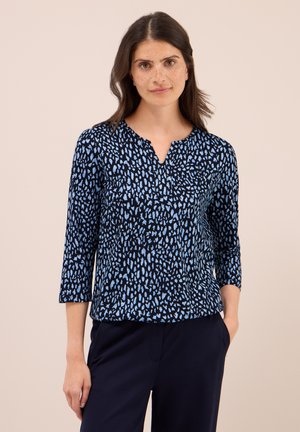 Femme aux longs cheveux noirs portant un haut à manches longues noir et bleu à motifs et un pantalon foncé, se tenant avec une main dans la poche.