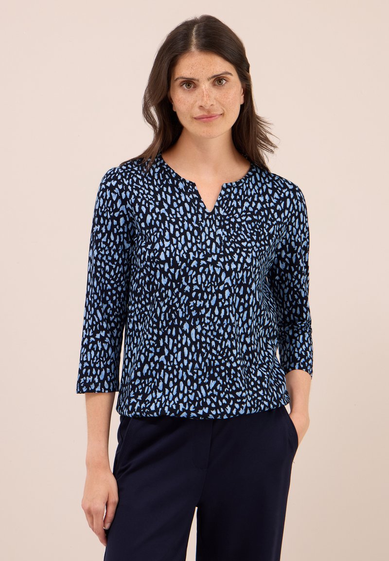 Mujer con cabello largo y oscuro, vistiendo una blusa de manga larga negra y azul con un patrón, y pantalones oscuros, de pie con una mano en el bolsillo.