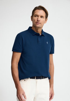 Mann trägt ein marineblaues kurzärmeliges Poloshirt mit einem kleinen weißen gestickten Logo, kombiniert mit weißen Hosen und einem schwarzen Gürtel.