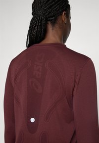 Persona con cabello trenzado que lleva una camiseta de manga larga de color rojo oscuro con patrones texturizados sutiles y un logotipo tenue en la parte posterior.