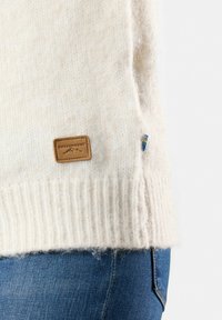 Crème-kleurige gebreide trui met ribbels aan de mouwen, voorzien van een leren label en een zij-accent in het blauw, gemaakt van zachte, gestructureerde stof.