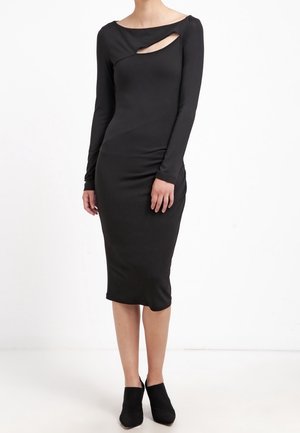 Vrouw die een aansluitende zwarte midi-jurk draagt met lange mouwen en een asymmetrische uitsnijding nabij de halslijn, gecombineerd met zwarte enkellaarzen.