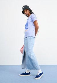 Light purple graphic T-shirt with blue design, paired with light blue denim maxi skirt. Pink handbag and blue sneakers complete the look.