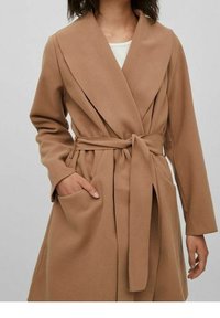 Long manteau cache-cœur marron avec un col profond, une ceinture nouée et des poches latérales. Fabriqué en tissu doux avec une texture lisse.