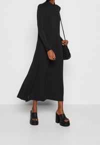 Musta pitkähiha dressi, jossa on korkea kaula-aukko ja sivurei'ät. Yhdistettynä jykeviin mustiin korokepohjakenkiin ja texturoituun mustaan käsilaukkuun.