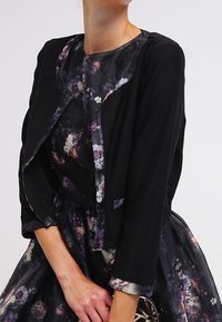 Svart lättviktsjacka över en blommönstrad klänning med figursydd överdel, svängig kjol och trekvartsärmar. Slät tygtextur.