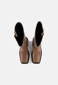 Botine din piele maro, cu vârf pătrat, catarame decorative și talpă din cauciuc negru contrastant. Textură netedă cu căptușeală interioară.