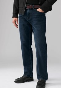 Jeans de mezclilla azul oscuro con corte recto, que presentan costuras en contraste y un cinturón marrón. Llevados con zapatos negros tipo slip-on.
