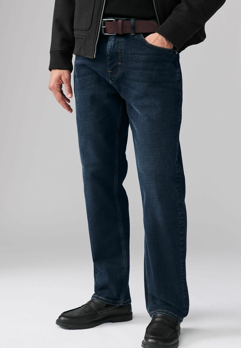 Jeans de mezclilla azul oscuro con corte recto, que presentan costuras en contraste y un cinturón marrón. Llevados con zapatos negros tipo slip-on.