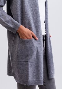 Grijze gebreide cardigan met lange mouwen, open voorkant, ribgebreide manchetten en twee voorzakken. Zachte textuur, casual ontwerp, kniehoogte.