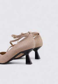 Zapatos de tacón alto de ante beige con correas ajustables en el tobillo y un tacón negro acampanado. El diseño presenta una punta redondeada y una textura suave.