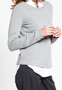 Kobieta w jasnoszarej, długosleeve swetrze na białej koszuli z kołnierzykiem i ciemnych spodniach, stoi z dłońmi złożonymi.