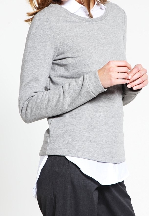 Kobieta w jasnoszarej, długosleeve swetrze na białej koszuli z kołnierzykiem i ciemnych spodniach, stoi z dłońmi złożonymi.