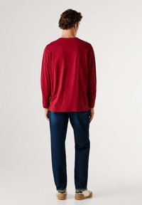 Maglietta rossa a maniche lunghe con scollo rotondo, vestibilità comoda e materiale in cotone, abbinata a jeans blu scuro e sneaker casual.