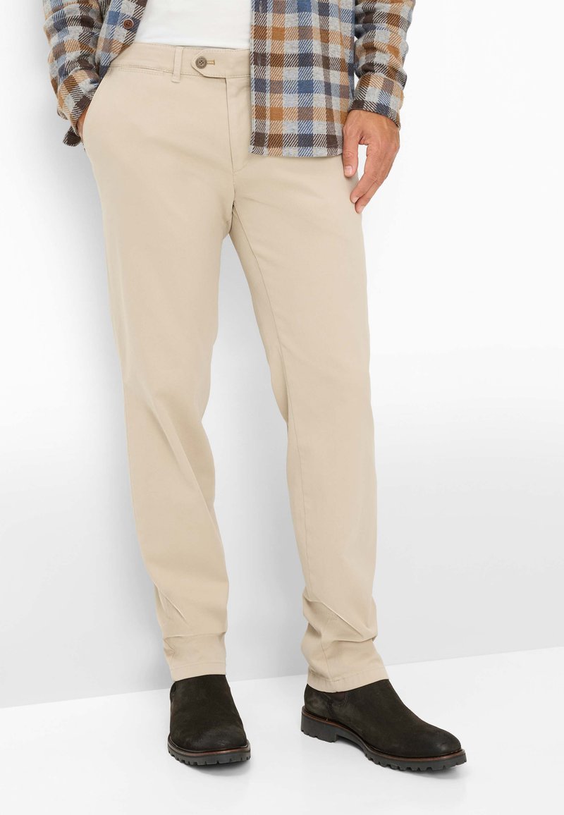 Pantaloni in cotone beige con taglio slim, caratterizzati da una tasca frontale, chiusura con bottone e abbinati a stivali alla caviglia marrone scuro.