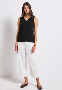 Czarny top bez rękawów z dekoltem w serek, w połączeniu z białymi spodniami o szerokich nogawkach. Modelka stoi boso, prezentując czarne plecione buty oraz złotą bransoletkę.