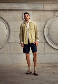 Svetlo olivová ľanová košeľa, biela košeľa s gombíkmi pod ňou, námornícke šortky, béžové slip-on topánky, stojí proti textúrovanej šedej stene s kruhovými detailmi.