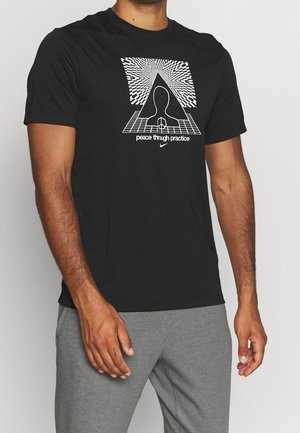 Homme portant un t-shirt noir avec un motif géométrique blanc, une silhouette, le texte "paix par la pratique" et un petit logo Nike, associé à un pantalon gris.