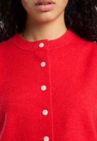 Red knitted cardigan featuring a round neckline, button closure, and four white buttons. The fabric has a soft texture.