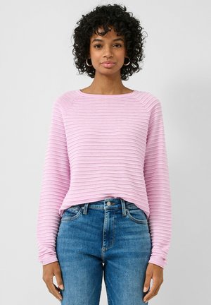 Femme aux cheveux bouclés portant des boucles d’oreilles créoles, un haut long rose côtelé et un jean bleu, debout devant un fond uni.