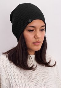Black ribbed knit beanie featuring a small logo tag, worn by a person with long dark hair, paired with a white textured sweater.