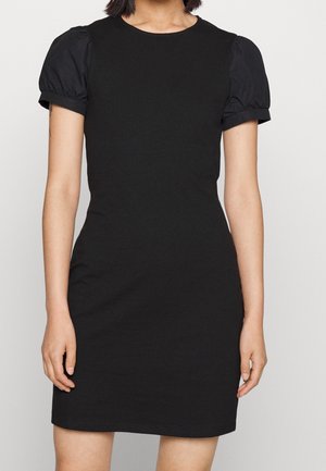 Kobieta w dopasowanej czarnej sukience z krótkimi bufkami i okrągłym dekoltem, stojąca na jednolitym jasnym tle.