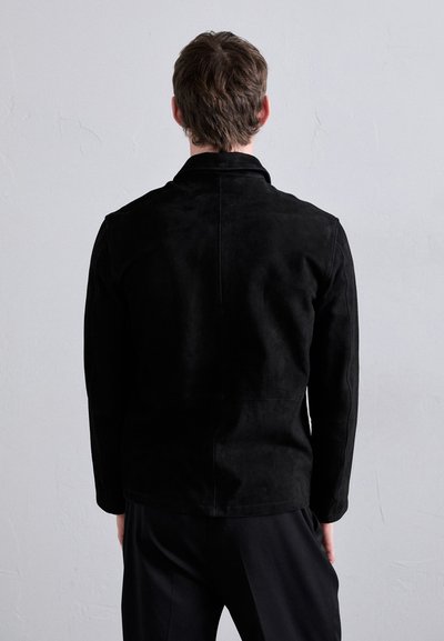 Veste en daim noir avec un col, des manches longues et une coupe droite. Texture lisse avec un design minimaliste et sans quincaillerie visible.