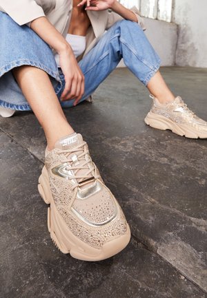 Person sitzt auf dem Boden und trägt hellblaue Jeans und beige klobige Sneaker, die mit Strasssteinen und metallischen Akzenten verziert sind.