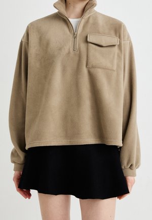 Person trägt beige Fleece-Pullover mit Viertelreißverschluss und Brusttasche, kombiniert mit einem kurzen schwarzen Rock vor einfachem Hintergrund.