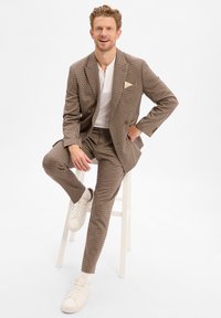 Brązowy garnitur w wzór houndstooth z dopasowaną marynarką i spodniami, w zestawieniu z białym polo i kremowymi sneakersami. Warto zauważyć detal w postaci chusteczki w kieszeni.
